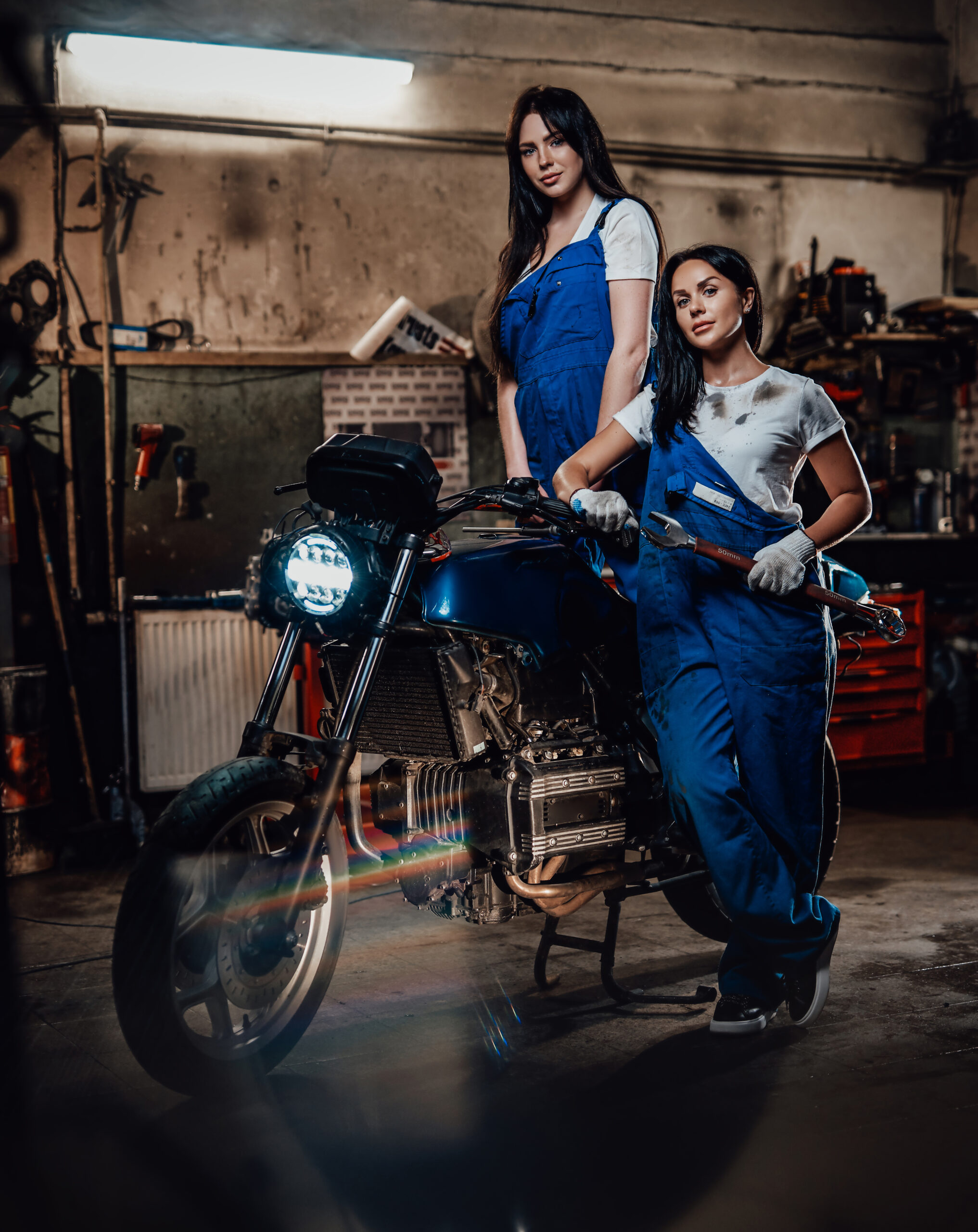 Two hot brunette women in blue overalls posing next to a custom bobber in authentic workshop garage